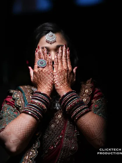An artistic bridal portrait focusing on the intricate henna on the bride's hands as she covers her face, with dramatic lighting highlighting her jewelry.