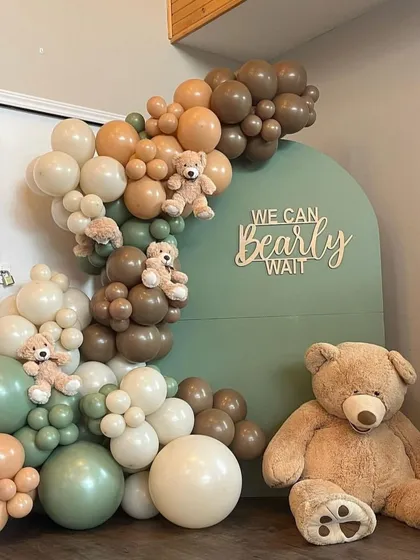 A charming 'We can Bearly Wait' baby shower theme with an earthy color scheme. The sage green backdrop is adorned with a balloon garland in brown, tan, and cream, and decorated with several small teddy bears.