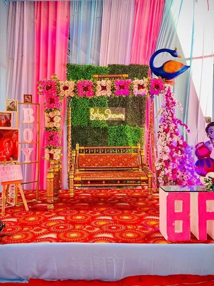 A baby shower stage with a traditional swing, decorated with pink and blue elements and Radha-Krishna cutouts.