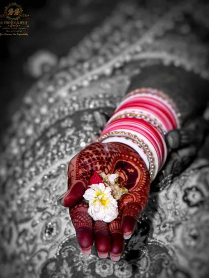 An artistic black and white photo with selective color, focusing on the bride's hand with intricate mehndi holding a small flower. This highlights the delicate details of the wedding rituals.