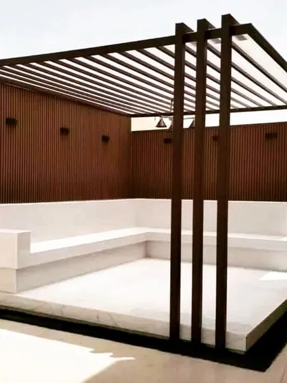 This pergola features a dark metal frame and built-in, multi-level concrete seating. The back wall is clad in vertical wood-style panels, offering a clean, minimalist, and durable solution.