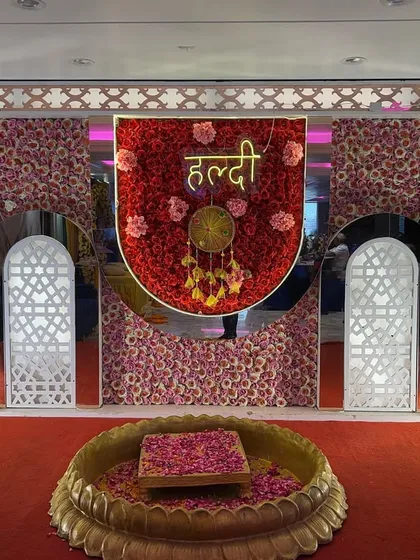A grand and royal Haldi setup featuring a backdrop made entirely of red, pink, and yellow flowers, with decorative arches and a central lotus bowl.