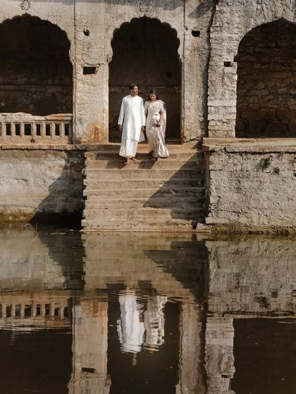 A serene moment captured at a historic stepwell, where the couple's reflection is perfectly mirrored in the calm water. Dressed in white, their presence adds a timeless elegance to the ancient stone architecture.