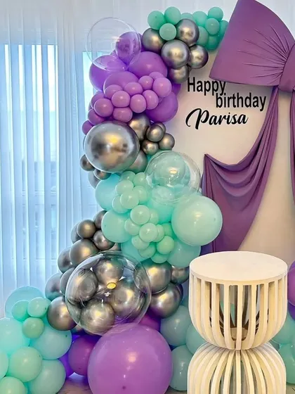 A luxurious birthday setup featuring a large purple bow and drapery on a white backdrop. The scene is framed by a balloon garland in mint green, purple, and silver for a unique and elegant color combination.