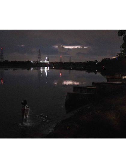 A person takes a spiritual bath in the Yamuna River before sunrise. The lights from the distant industrial area reflect on the water, creating a moody and atmospheric scene.