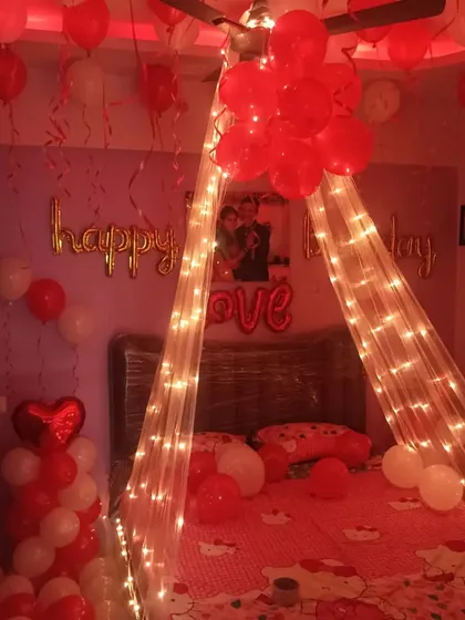 A romantic birthday surprise with a canopy of net and fairy lights over the bed, surrounded by red and white balloons.