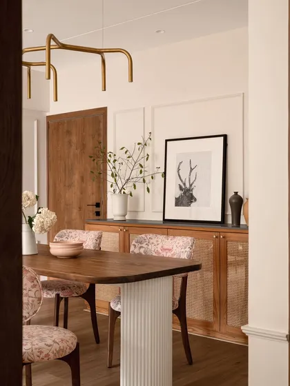 The dining area in "Apartment 222" showcases a blend of modern and traditional elements. A minimal gold metal light fixture hangs above the wooden table, while the cane-fronted sideboard and framed art add layers of texture and personality.