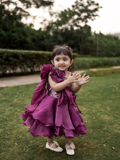 A little girl in a frilly purple dress claps her hands with a happy expression during her outdoor photoshoot.