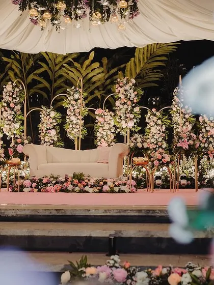 A perspective of the reception stage from a distance, framed by foreground flowers. This view shows the depth and dimension of the floral arrangement, making the stage feel like a secret garden.