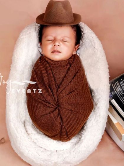 A touch of character for your little one's first photoshoot. This newborn is styled as a little scholar or cowboy, swaddled in a brown wrap with a matching fedora, resting comfortably in a soft, plush pod.