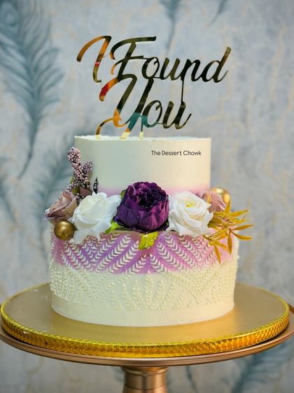 An elegant two-tier engagement cake with a unique stencil pattern on the bottom tier. The design is finished with beautiful flowers and a custom "I Found You" topper.