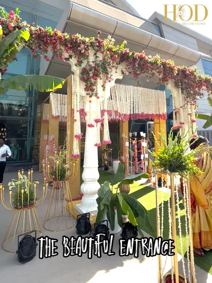 A magnificent outdoor entrance combining traditional white pillars with a canopy of bougainvillea and hanging jasmine flowers, creating a classic South Indian wedding feel.