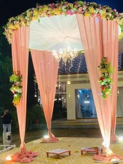 A beautiful and romantic outdoor wedding mandap. The design features a circular structure with delicate peach-colored drapes, a classic chandelier for evening ceremonies, and lush floral arrangements, creating a perfect setting for your vows.
