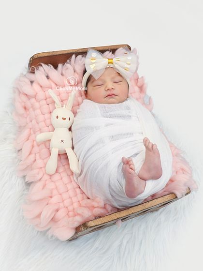 This little girl looks angelic in a simple white swaddle and a big bow headband, resting on a soft pink blanket with her bunny friend.