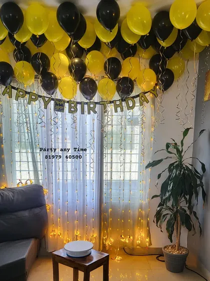 A birthday surprise setup with black and yellow helium balloons, a 'Happy Birthday' banner, and fairy lights.