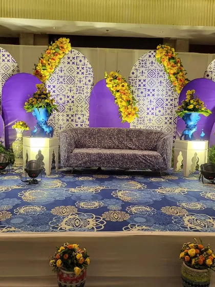 The main seating stage for a blue pottery themed Haldi. The backdrop features printed jaali patterns and purple arches, complemented by blue vases and yellow marigolds for a striking color contrast.