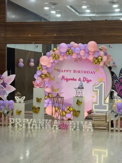 A fairy and butterfly theme for a joint first birthday celebration. The decor features a soft pink backdrop, pastel and gold balloon arrangements, and marquee letters for both names.
