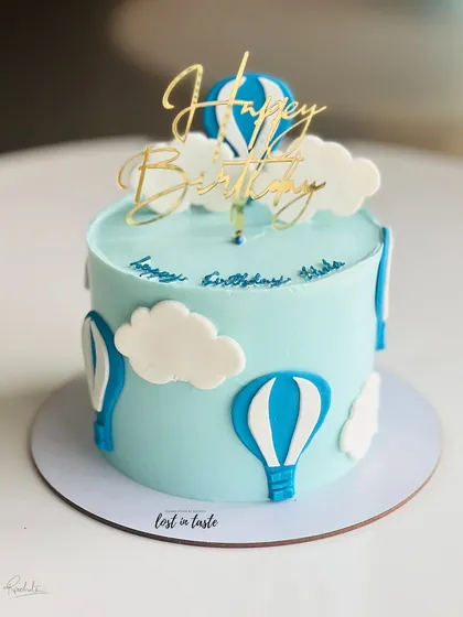 A whimsical hot air balloon themed cake. The light blue buttercream sky, fluffy clouds, and a golden "Happy Birthday" topper make it a delightful choice for a child's celebration.