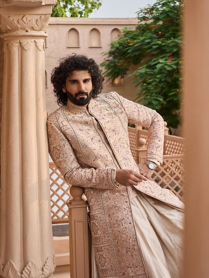 This peach-toned sherwani offers a warm and contemporary option for the groom. The intricate, all-over embroidery adds a layer of opulence, seen here in a relaxed, confident pose.