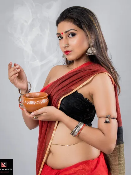 A portrait capturing a moment of quiet tradition. The model, dressed in a red saree, holds a steaming earthen cup, blending cultural elements with a soft, ethereal mood in a studio setting.