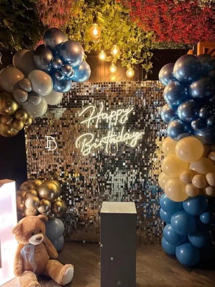A first birthday setup combining a silver sequin wall with a teddy bear theme. The decor uses blue and gold balloons and includes a cute teddy bear sitting next to the display.