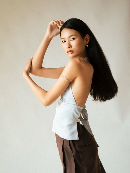 A minimalist studio shot focusing on form and silhouette. The model's elegant pose and the simple, clean background draw attention to the backless design of the halter top and the flow of her hair.