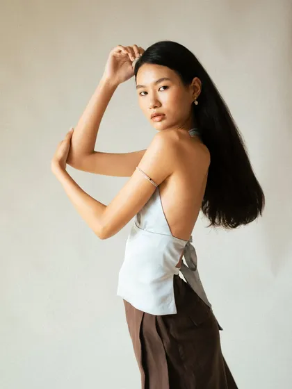 A minimalist studio shot focusing on form and silhouette. The model's elegant pose and the simple, clean background draw attention to the backless design of the halter top and the flow of her hair.