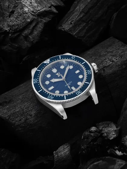 A macro shot of a blue-faced watch on a bed of black charcoal. The contrast in texture and color makes the watch's details pop, giving it a rugged yet sophisticated look.