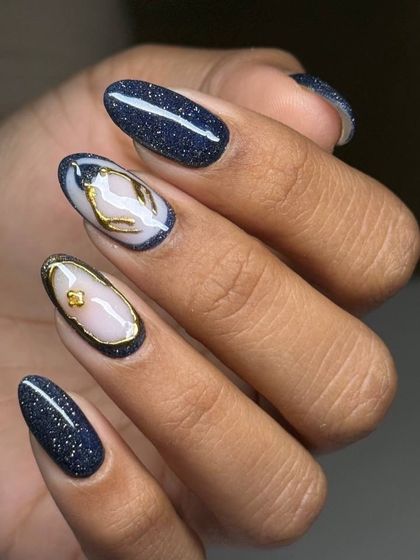 An elegant and artistic design featuring a deep, sparkling navy base with abstract gold and milky white details on two accent nails, resembling a precious stone.