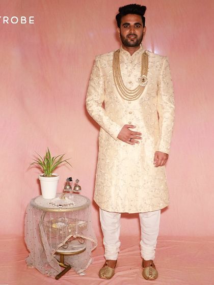 For a truly royal feel, this golden-beige brocade sherwani is a standout choice. Paired with a multi-layered pearl and ruby mala, it’s designed for a groom who wants to shine.