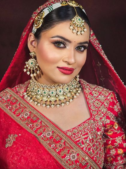 A close-up portrait of a bride in a classic red lehenga. The makeup features a flawless base, defined eyes, and a bold red lip, creating a timeless and elegant look.