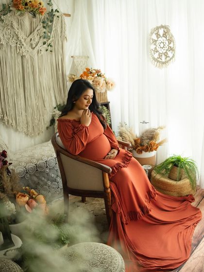 A beautiful seated portrait in a rich, rust-colored gown. The autumn tones of the dress and the surrounding boho decor create a warm and cozy atmosphere.