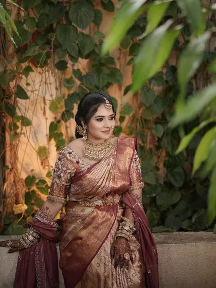 A bride in a wine-colored saree, posing in a beautiful garden setting.