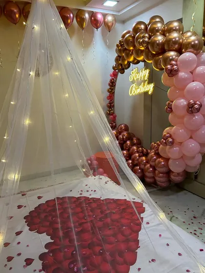 A close-up of the bed decoration from the romantic room setup. It features a heart made of red rose petals on the bed sheet, under a canopy illuminated with warm fairy lights.