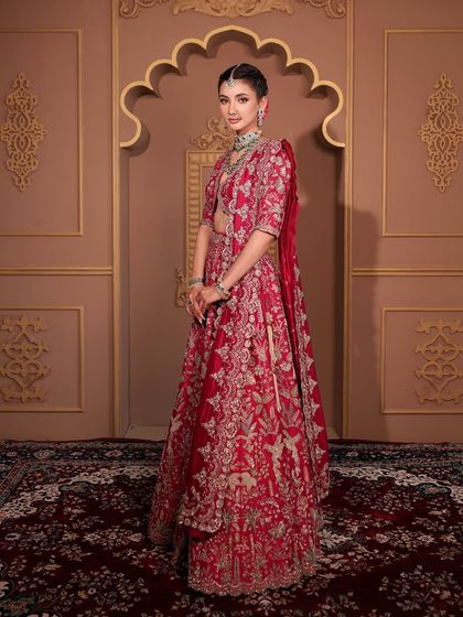 A full-length portrait of a bride in a magnificent red lehenga. Her makeup is kept elegant and understated to balance the grandeur of her outfit, focusing on a flawless complexion and defined features.