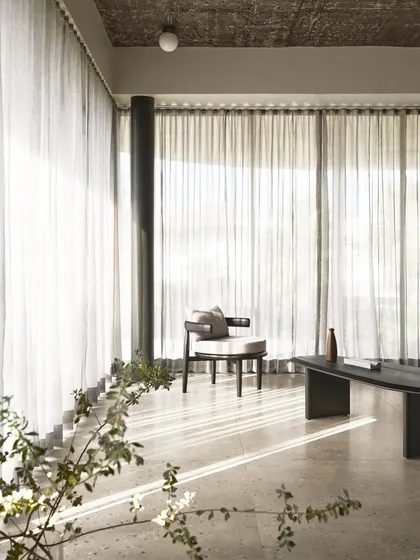 Sunlight streams into the living area of Pavilion House, highlighting the delicate sheer curtains and the simple, elegant form of the curated armchair. The space is a meditation on light, shadow, and minimalist comfort.