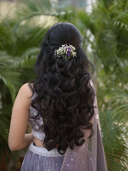 An elegant half-up, half-down hairstyle for an engagement. Soft curls cascade down, while the top is adorned with a delicate spray of baby's breath and a hint of purple flowers to match her outfit.