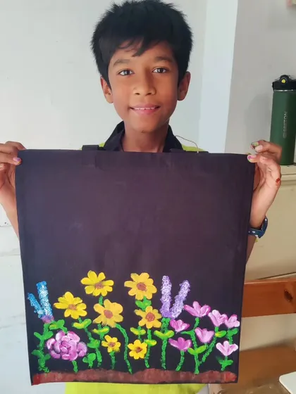 A young boy proudly holds up a black tote bag he painted with a beautiful garden of yellow, pink, and purple flowers. This was part of a pre-mela workshop at a local school.