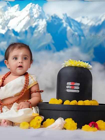 A toddler version of the Bala Shiva theme, with the baby sitting up next to a cake-like Shivling against a backdrop of the Himalayas.