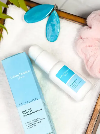 This flatlay shows a moisturizer next to its box on a wooden tray with a soft towel and accessories. The overhead shot gives a clear view of the product and its packaging in a relaxed, everyday context.