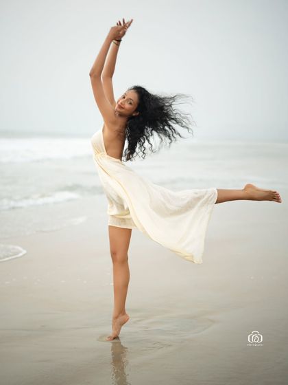 A dynamic and graceful dance-inspired pose on the beach. The model's energy and the movement of the white dress create a powerful and expressive fashion photograph.