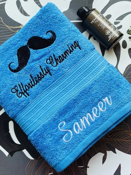 An 'Effortlessly Charming' personalised towel in bright blue, embroidered with a mustache motif and the name 'Sameer'. A great gift for him.