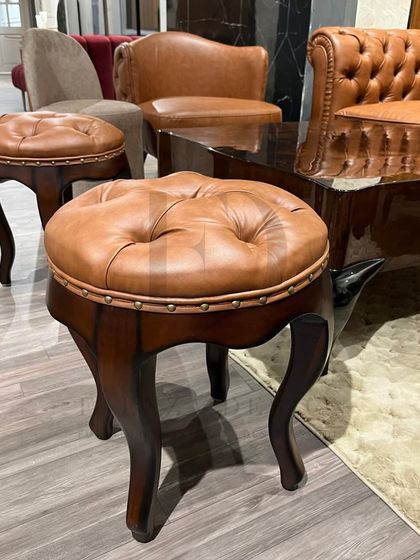 A classic living room setup featuring a brown leather Chesterfield sofa, matching tufted stools with cabriole legs, and complementary armchairs. This collection exudes a warm, traditional, and clubby feel.