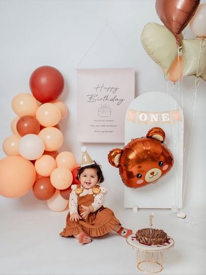 A happy birthday girl sits next to her cake in a studio setup decorated with a balloon arch and a cute teddy bear theme.