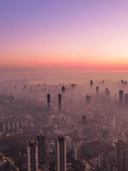 A wide drone shot of Mumbai enveloped in a sea of smog, with the city's skyscrapers barely visible through the haze at sunset.