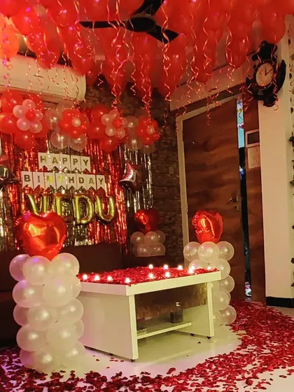 A full living room birthday decoration in a classic red and white theme. The setup includes a shimmery foil curtain backdrop, balloon pillars, and a candle-lit center table covered in rose petals.