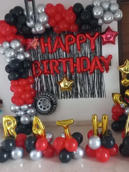 A Hot Wheels birthday backdrop for Pratha. This home setup features a balloon arch with car-themed foil balloons, a tire prop, and the name spelled out in gold letters.