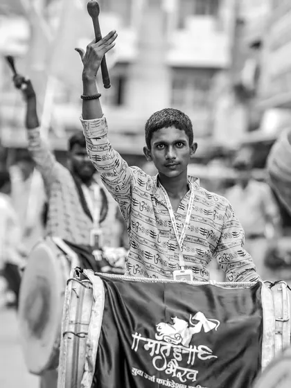 A young dhol player raises his sticks, his expression one of proud participation.