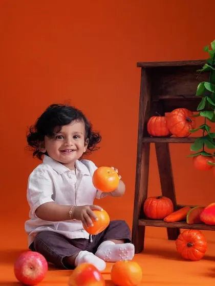 A happy toddler sits amidst a vibrant orange-themed setup, playing with oranges and pumpkins against a matching backdrop.