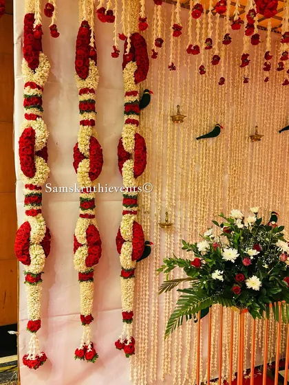 A close-up of unique, knotted flower garlands made from red and white petals. Small bird motifs and traditional diyas are placed on the backdrop to complete the look.